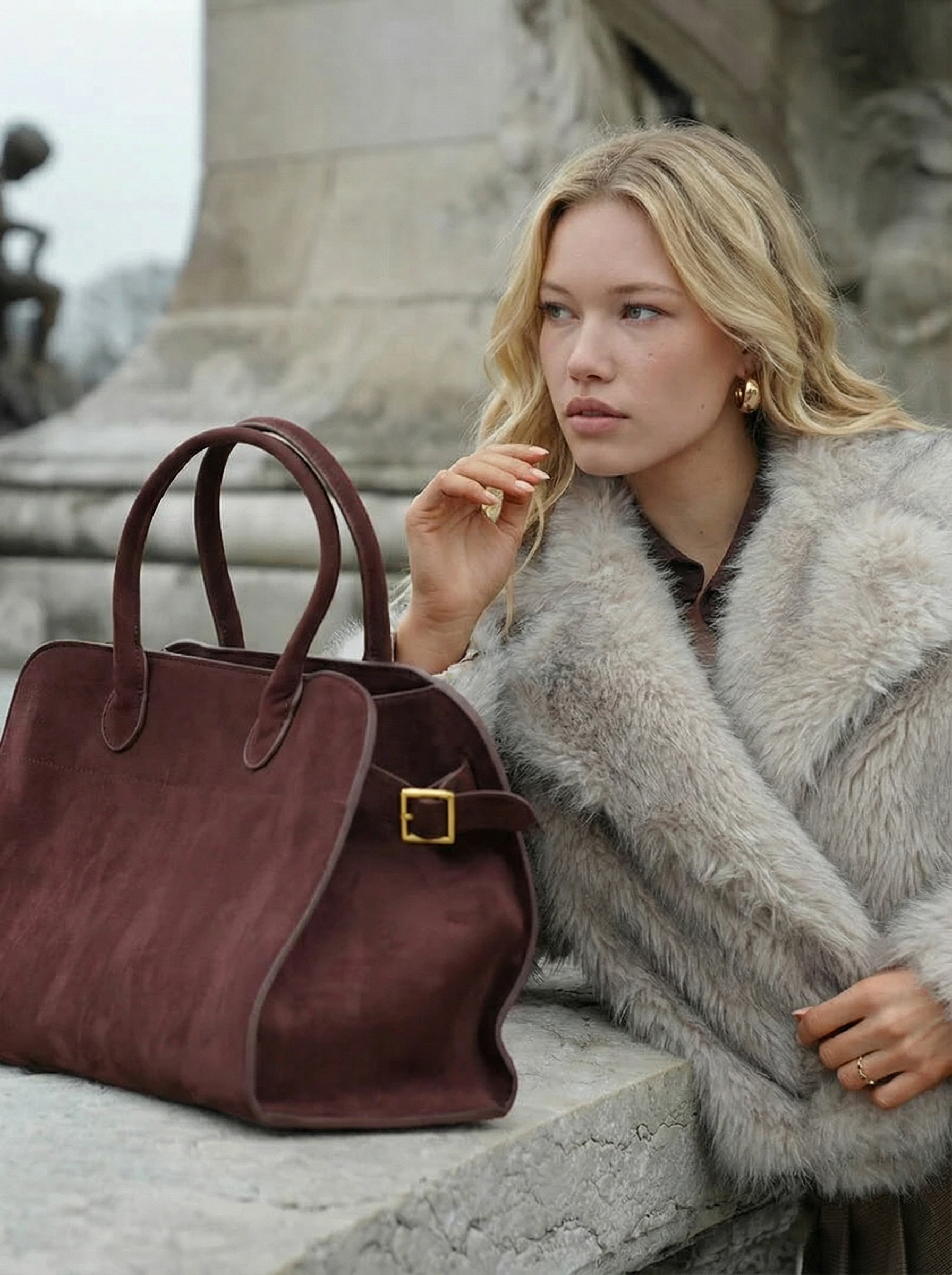 Woman in a fur coat with a brown handbag sitting on a stone bench.